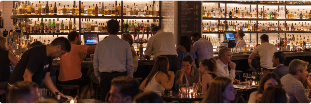 Lively bar scene with patrons seated and standing around a well-stocked counter, bartenders serving drinks, and dim lighting creating a warm ambiance—used to illustrate AML training context for hospitality venues.